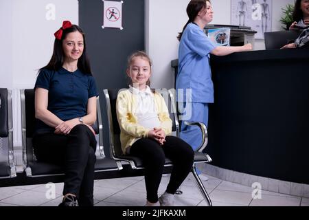 Smiling woman and her child attending pediatrician appointment in private clinic to do routine checkup. Mother and young daughter waiting for pediatric doctor while nurse is talking with receptionist. Stock Photo