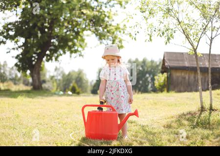 A little cute baby girl 3-4 years old in a dress watering the plants from a watering can in the garden. Kids having fun gardening on a bright sunny Stock Photo