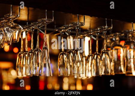 Empty clean upside-down crystal wine glasses hanging from the bar in luxury restaurant. Selective focus blurred colourful background Stock Photo