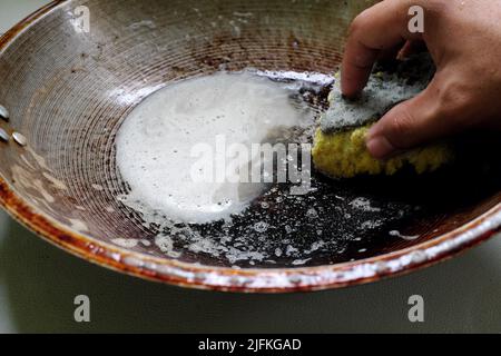 Human hand scrubbing or cleaning a burnt, scorched and greasy pan or wok on kitchen sink. Stock Photo