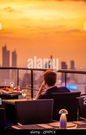 Young man with glass of wine rest at luxury rooftop restaurant watching orange sky sunset. Male with cocktail drink at sky bar terrace looking at Stock Photo