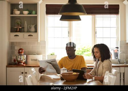 Young multiethnic couple catching on news during breakfast in their kitchen Stock Photo