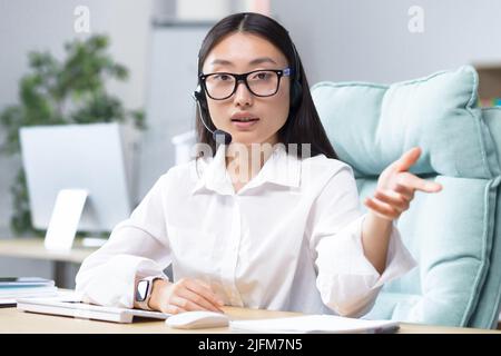 Beautiful Asian female bank support technician looking into camera and talking on video call, using headset, explaining information to customers, helpline. Stock Photo