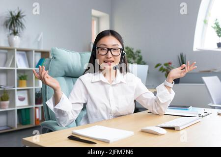 Beautiful Asian female bank support technician looking into camera and talking on video call, using headset, explaining information to customers, helpline. Stock Photo