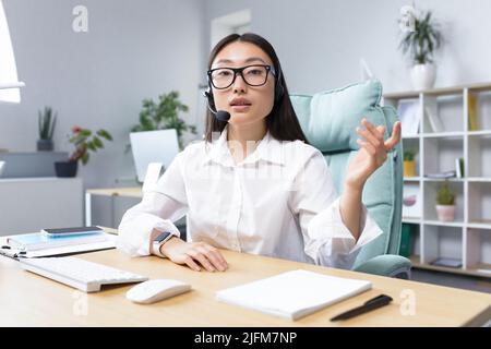 Beautiful Asian female bank support technician looking into camera and talking on video call, using headset, explaining information to customers, helpline. Stock Photo