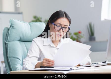 Asian business woman behind paper work, tired and frustrated, working in office, female employee in bra looking at documents and financial reports. Stock Photo