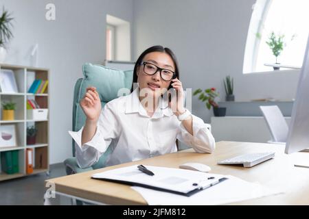 Beautiful asian office worker secretary working in modern office talking on the phone smiling and looking at camera. Stock Photo