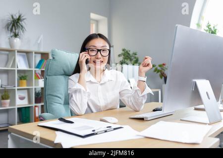 Beautiful asian office worker secretary working in modern office talking on the phone smiling and looking at camera. Stock Photo