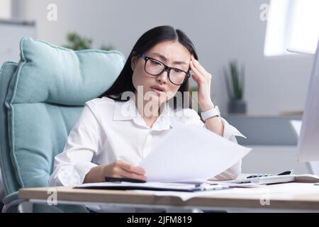 Young Asian woman working on paper in office, businesswoman anxiously examines documents and financial reports. Stock Photo