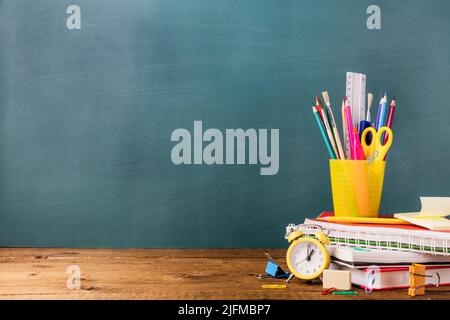 Many different various school, office supplies and stationery. Back to school concept. Notebook, book, pen, pencil, scissors, alarm clock, clock, sticker stand on a wooden table on a green background. Stock Photo