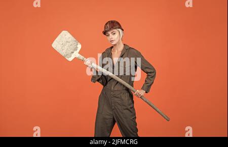 woman hard worker in protective helmet and boilersuit hold shovel on orange background, labour day Stock Photo