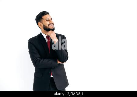 Positive elegant influential arabian or indian man in formal suit, standing over isolated white background, looking away, thinking, planning, dreaming, smiles happily. Copy-space for your presentation Stock Photo