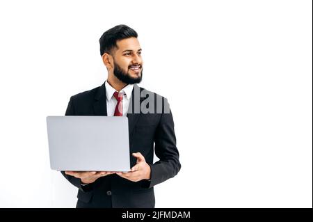 Positive handsome elegant indian or arabian young man in business suit, male executive, holding open laptop in hands, looking aside, standing over isolated white background, smile friendly. Copy space Stock Photo