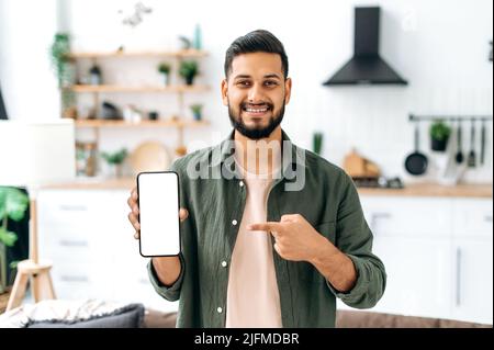 Positive handsome confident indian or arabian guy, stands in the living room against the background of the kitchen, shows a smartphone with a blank white mockup screen, points his finger at it, smiles Stock Photo