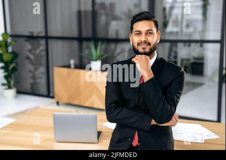 Successful attractive confident businessman of indian and arabian ethnicity, in a formal suit, business owner, ceo company, stands in his office near the workplace, looks at camera, smiles friendly Stock Photo