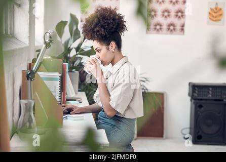Young focused mixed race businesswoman drinking a cup of coffee while working on a laptop at work. One hispanic female businessperson having a coffee Stock Photo