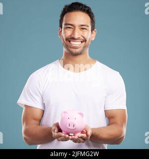 Handsome young mixed race man holding his pink piggybank while standing in studio isolated against a blue background. Hispanic male showing savings Stock Photo
