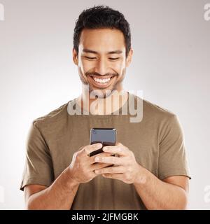 Handsome young mixed race man using his phone while standing in studio isolated against a grey background. Hispanic male sending a text message, using Stock Photo