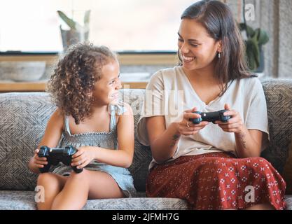 Hispanic mother and daughter playing video games together while sitting on the couch at home. Fun young mother and daughter using joysticks while Stock Photo