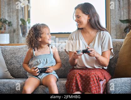 Hispanic mother and daughter playing video games together while sitting on the couch at home. Fun young mother and daughter using joysticks while Stock Photo