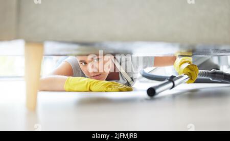 A mixed race domestic worker looking while lying on the floor and cleaning under the sofa. One mixed race female using a vacuum cleaner under a couch Stock Photo