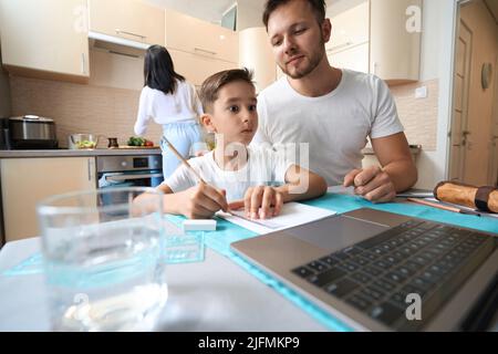 Man helping child with homework at laptop in kitchen Stock Photo