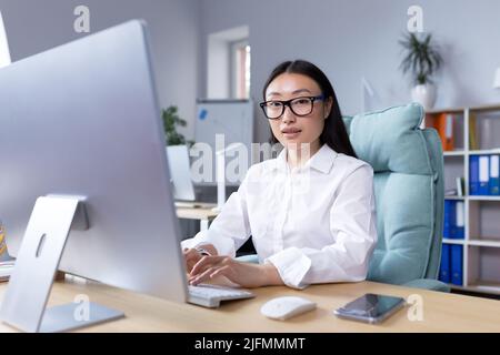 Portrait of successful office worker Asian woman, looking at camera smiling, business woman in glasses working and typing on computer. Stock Photo