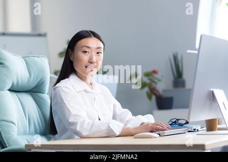 Beautiful Asian woman works at the computer in the office, looks at the camera and smiles, business woman in a white shirt typing on the keyboard. Stock Photo