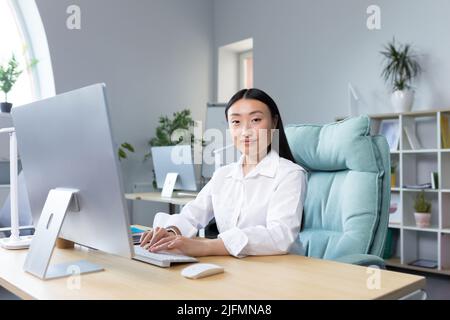 Beautiful Asian woman works at the computer in the office, looks at the camera and smiles, business woman in a white shirt typing on the keyboard. Stock Photo