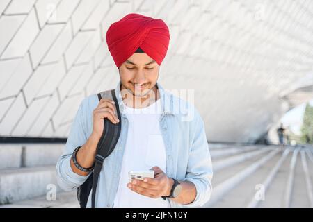 Indian male freelancer, student, entrepreneur with backpack wearing red national turban pagri using smartphone walking outdoors, hindu guy with phone in traditional headwrap texting, chatting online Stock Photo