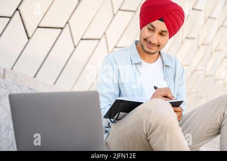 Modern Indian man in turban working with laptop outdoors. Hindu male in smart casual shirt watching webinar and writing down, taking notes with a pen, planning schedule Stock Photo