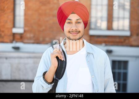 Inspired Indian male student in traditional turban with backpack outdoors, foreigner hindu guy proud to be university member, looking at the camera standing outdoors Stock Photo