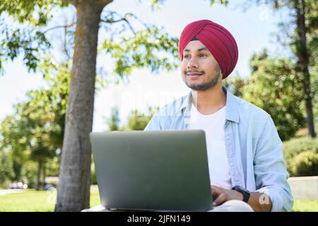 Smart inspired male Indian entrepreneur wearing red traditional headwrap turban pheta using laptop computer sitting on the lawn, hindu young man looking away dreamly, full of ambitions and energy Stock Photo