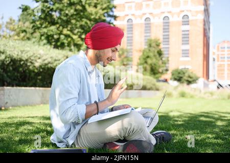 Male Indian student wearing red traditional headwrap turban pheta using laptop computer for video connection sitting on the lawn near campus, hindu young man waving, greeting classmates on distance Stock Photo