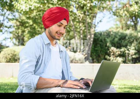 Optimistic ethnic man in national turban pagg using laptop sitting on the lawn in campus area, young indian male student watching webinars, educational courses, learning on the distance, typing emails Stock Photo