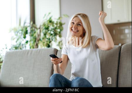 Leisure and entertainment concept. Positive excited caucasian blonde woman watching TV show or film, holding remote control, changes channels, while sitting on cozy sofa at home in living room, smiles Stock Photo
