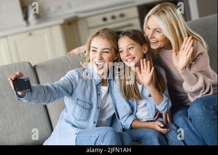 Three generations of Caucasian females. Happy grandmother, daughter and granddaughter, sitting on sofa in living room, having fun together, taking selfie posing for the camera, talking on a video call Stock Photo