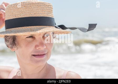 Slightly smiling gray haired woman holding boater from sea wind fluttering black ribbon on her hat against blurred background of sea with big waves. Stock Photo