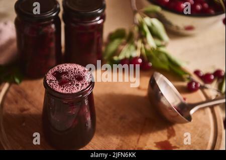 Close-up view of spilled poured freshly made cherry jam in sterilized glass jars and ladle on wooden board, on kitchen countertop. Homemade organic co Stock Photo