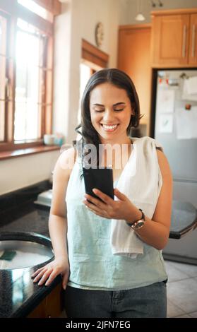Happy mixed race woman smiling while using smart phone at home. Woman reading text message or chatting on social media while busy with chores in the Stock Photo