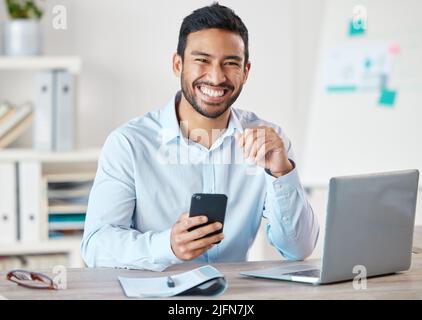 Portrait of a young handsome happy mixed race businessman using social media on his phone while working alone on a laptop in an office at work. One Stock Photo