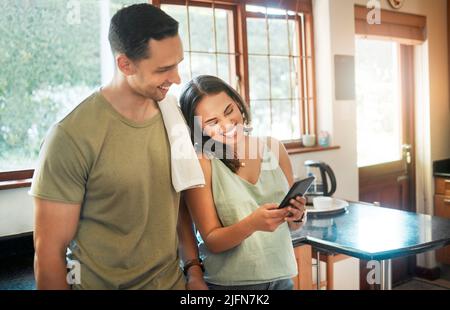 Young cheerful mixed race couple using a phone together in kitchen at home. Hispanic girlfriend showing her boyfriend a funny picture on her cellphone Stock Photo