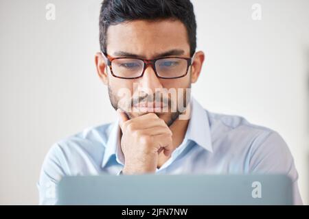 One focused mixed race businessman wearing glasses thinking of ideas while planning to brainstorm on a laptop in an office. Young motivated Stock Photo