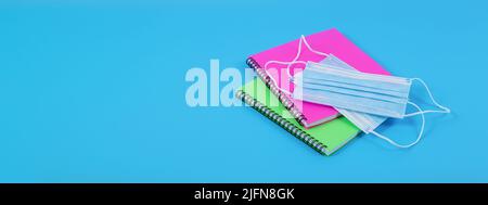 Pandemic back to school banner for website. Two colourful notebooks and blue protective face masks isolated on blue background, shot at angle. Stock Photo