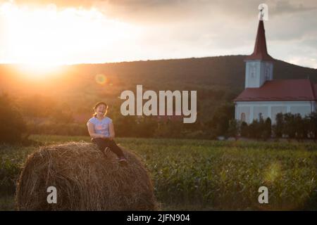 Caucasian girl having fun sitting on top of a golden hay bale on summer field near farm. Happy childhood and freedom concept. Rural countryside scenic Stock Photo