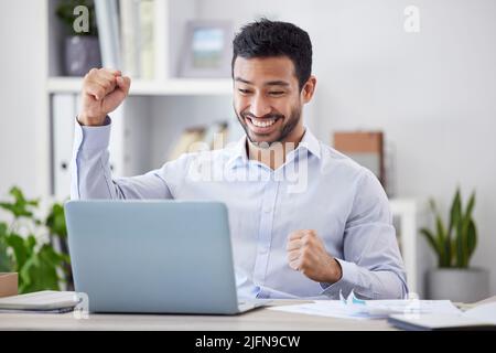 Young happy and excited mixed race businessman cheering with his fist working on a laptop sitting in an office at work. Hispanic businessperson Stock Photo
