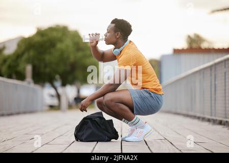 An African American man drinking water on a bridge. A man taking a break from exercising drinking from a bottle. A sporty guy focusing on health. Stock Photo