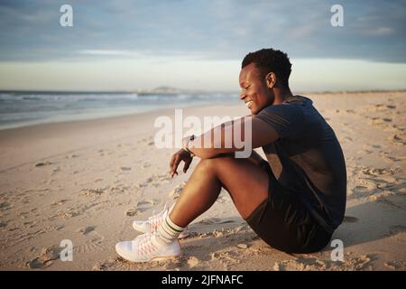 An african american muscular man looking happy and carefree while sitting at the beach. Black fit male cheerful relaxing on the sand on the beach Stock Photo