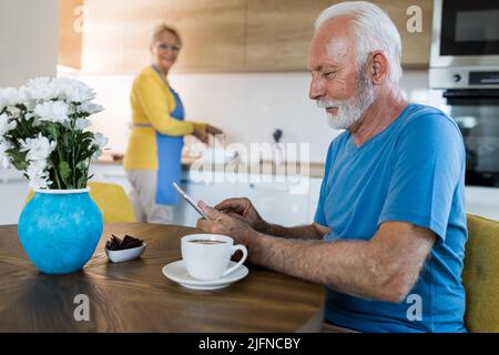 Senior man drinking coffee and looking at tablet at dining  table while woman preparing food in kitchen behind him Stock Photo