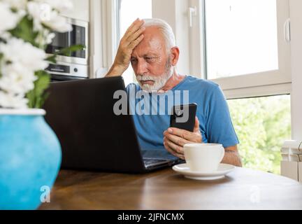 Upset senior man sitting at dining table at home holding mobile phone, and looking at laptop Stock Photo
